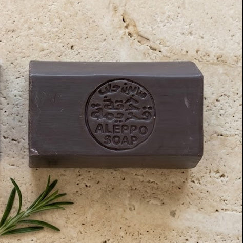 Two rectangular bars of Aleppo soap displayed on a textured beige stone slab. The left bar is in a purple paper sleeve with Arabic text and a small yellow square, and the right bar is dark gray with a circular embossed logo reading 'ALEHPPO SOAP'.