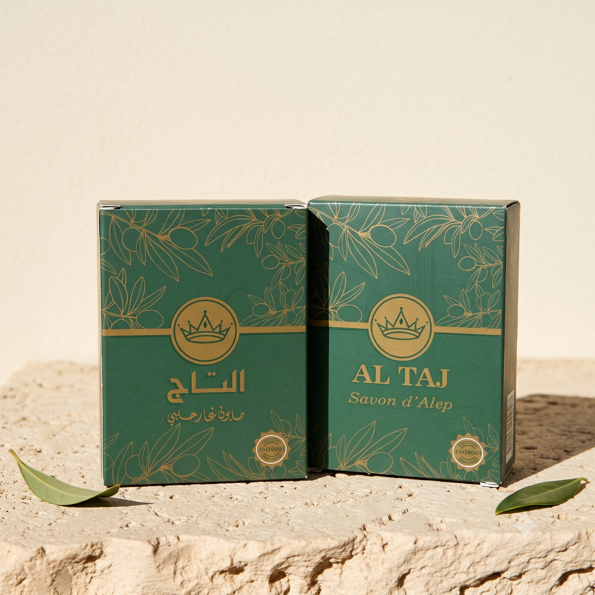 علبتان بلون أخضر وذهبي لصابون حلب العطري من التاج، على سطح حجري مع أوراق خضراء أمام خلفية بيج — صابون الغار الحلبي