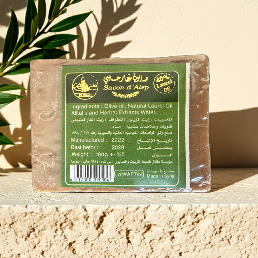 A rectangular bar of Savon d'Alep soap wrapped in clear plastic with a green and white label, displayed on a textured beige stone surface with leaf shadows cast across it — Aleppo laurel soap.