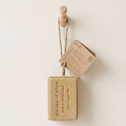 Aleppo Laurel Soap by Fansa — tan bar of Aleppo soap hanging by its rope beside a wooden surface, with dried leaves