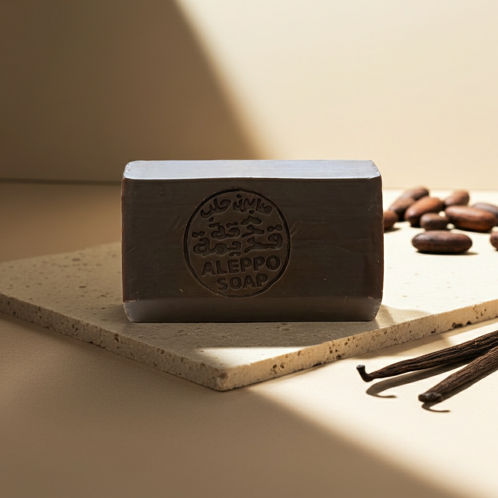 A rectangular bar of Aleppo soap in a dark brown color, embossed with a circular logo and Arabic text, resting on a textured stone slab with coffee beans and vanilla beans nearby.