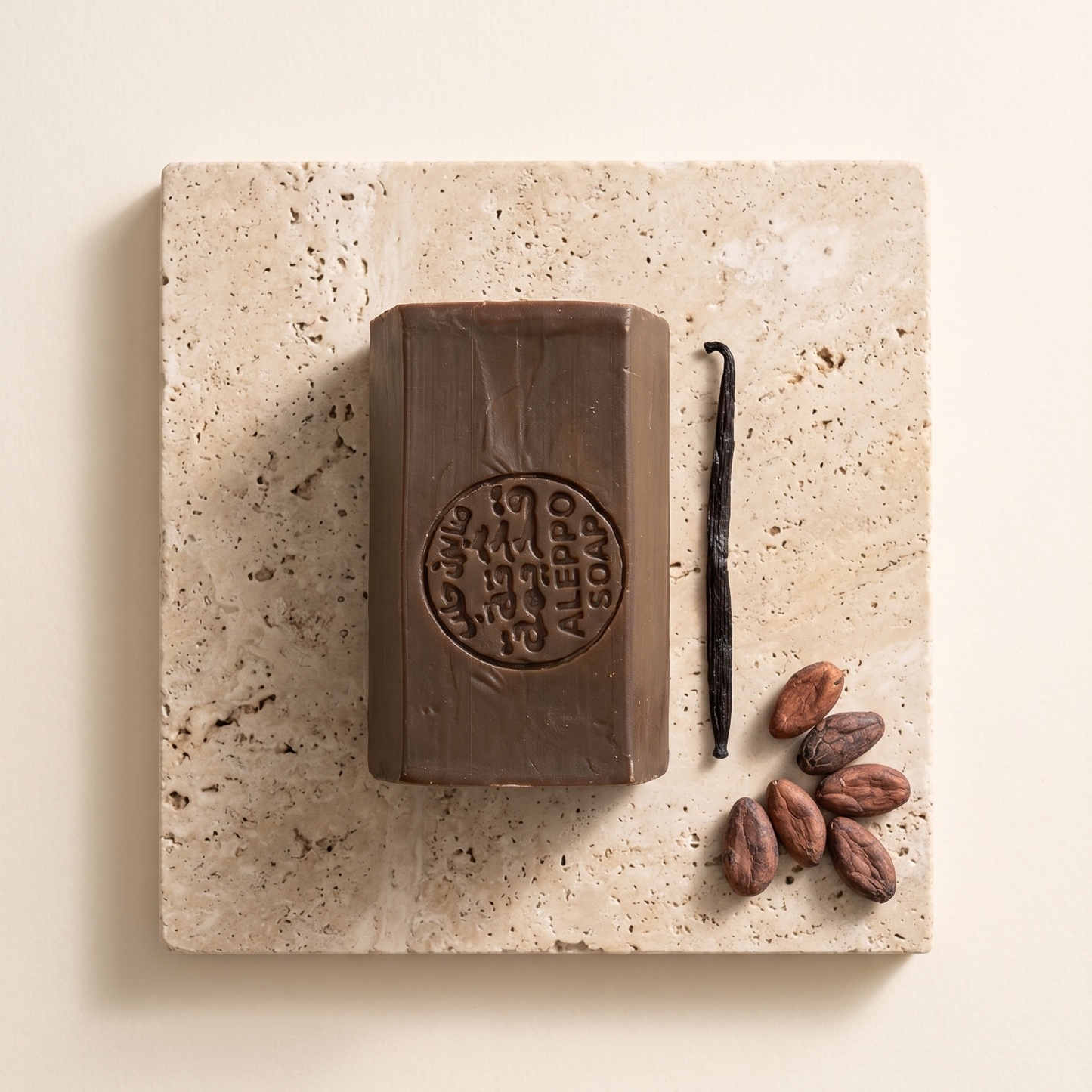 A rectangular bar of dark chocolate soap with an embossed circular logo, displayed on a square beige travertine stone slab alongside a vanilla bean and cocoa beans on a warm cream background.