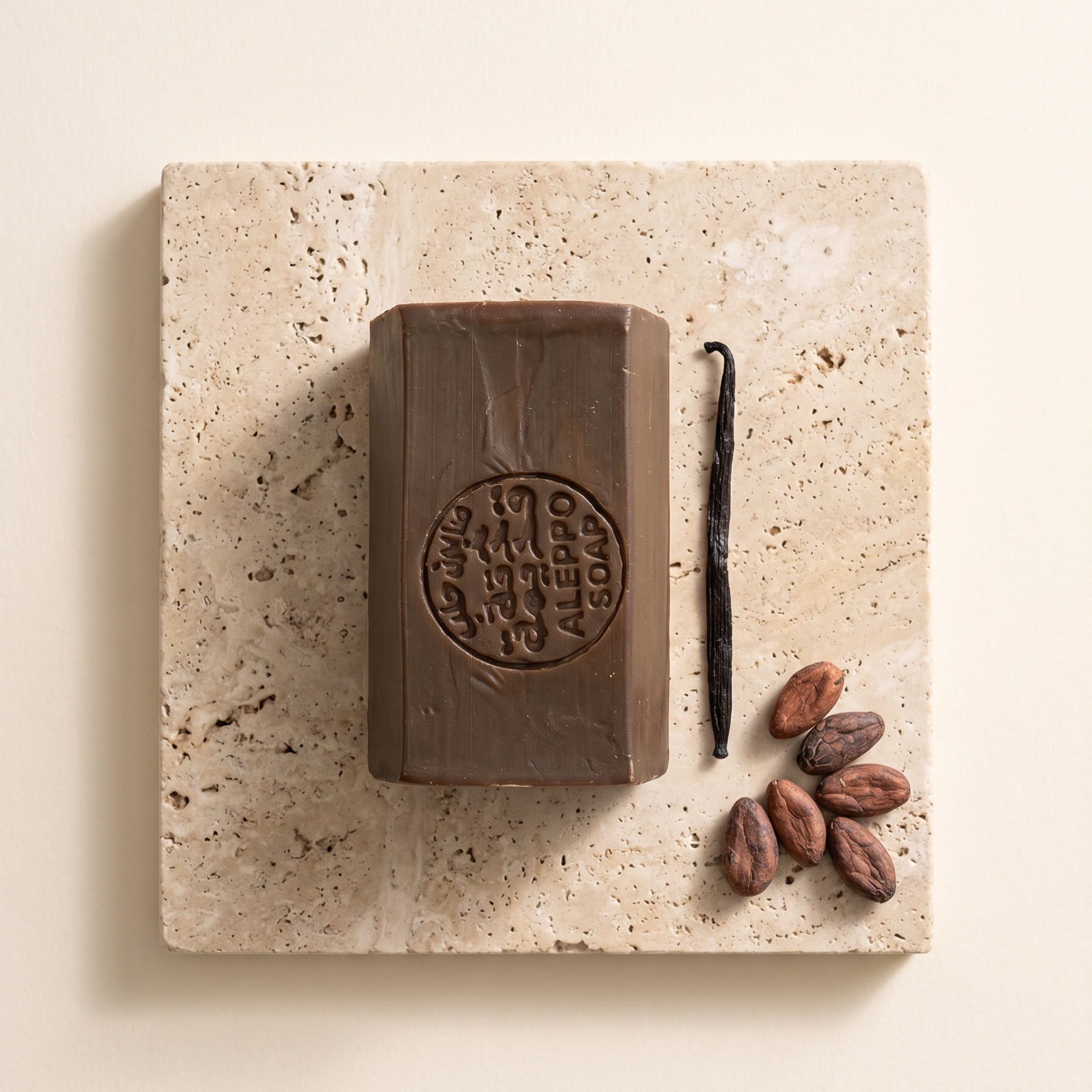 A rectangular bar of dark chocolate soap with an embossed circular logo, displayed on a square beige travertine stone slab alongside a vanilla bean and cocoa beans on a warm cream background.