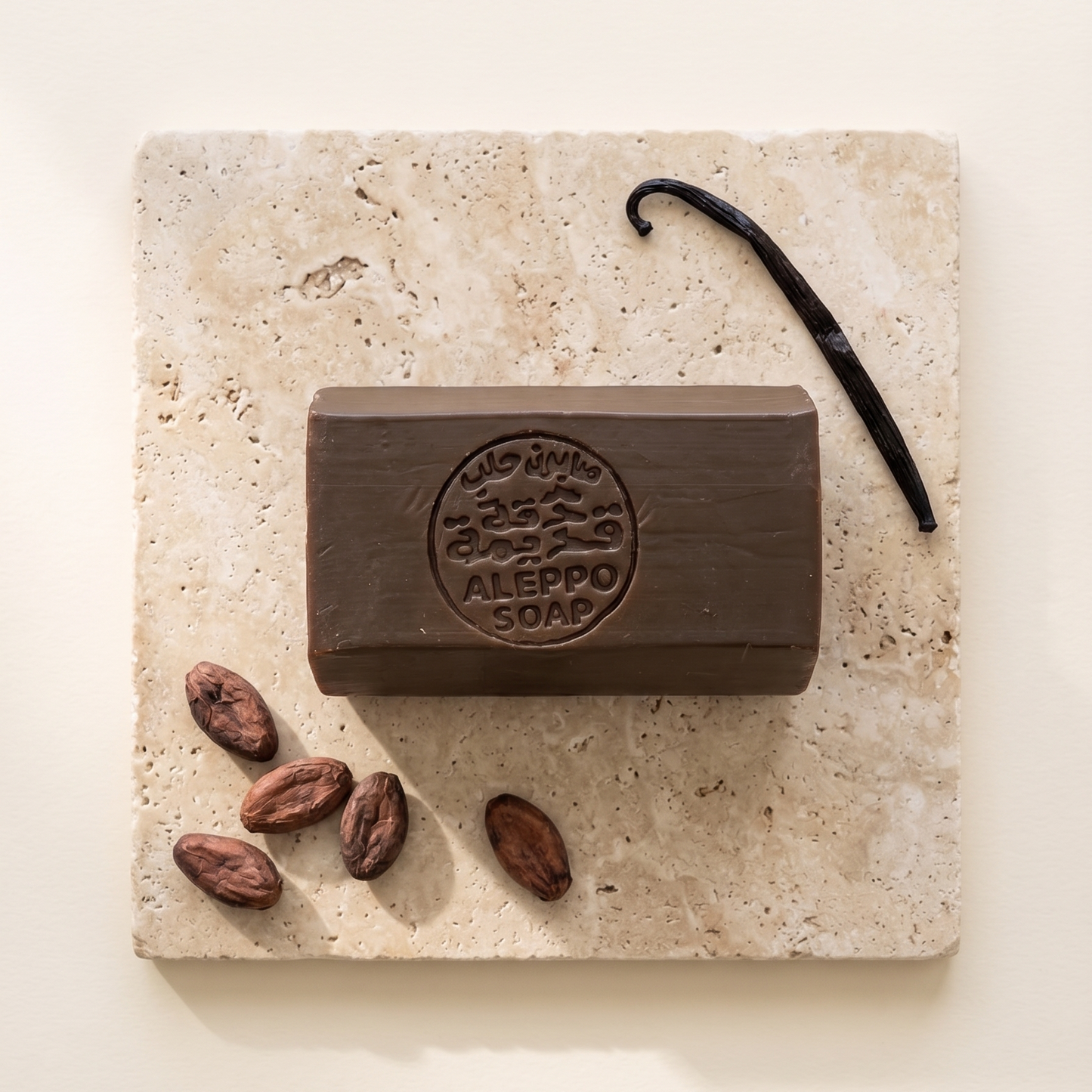 A rectangular bar of dark brown soap labeled 'MIO VINO ALEPPO SOAP' sits on a light beige stone slab, styled with cocoa beans and a vanilla bean on a warm cream background.