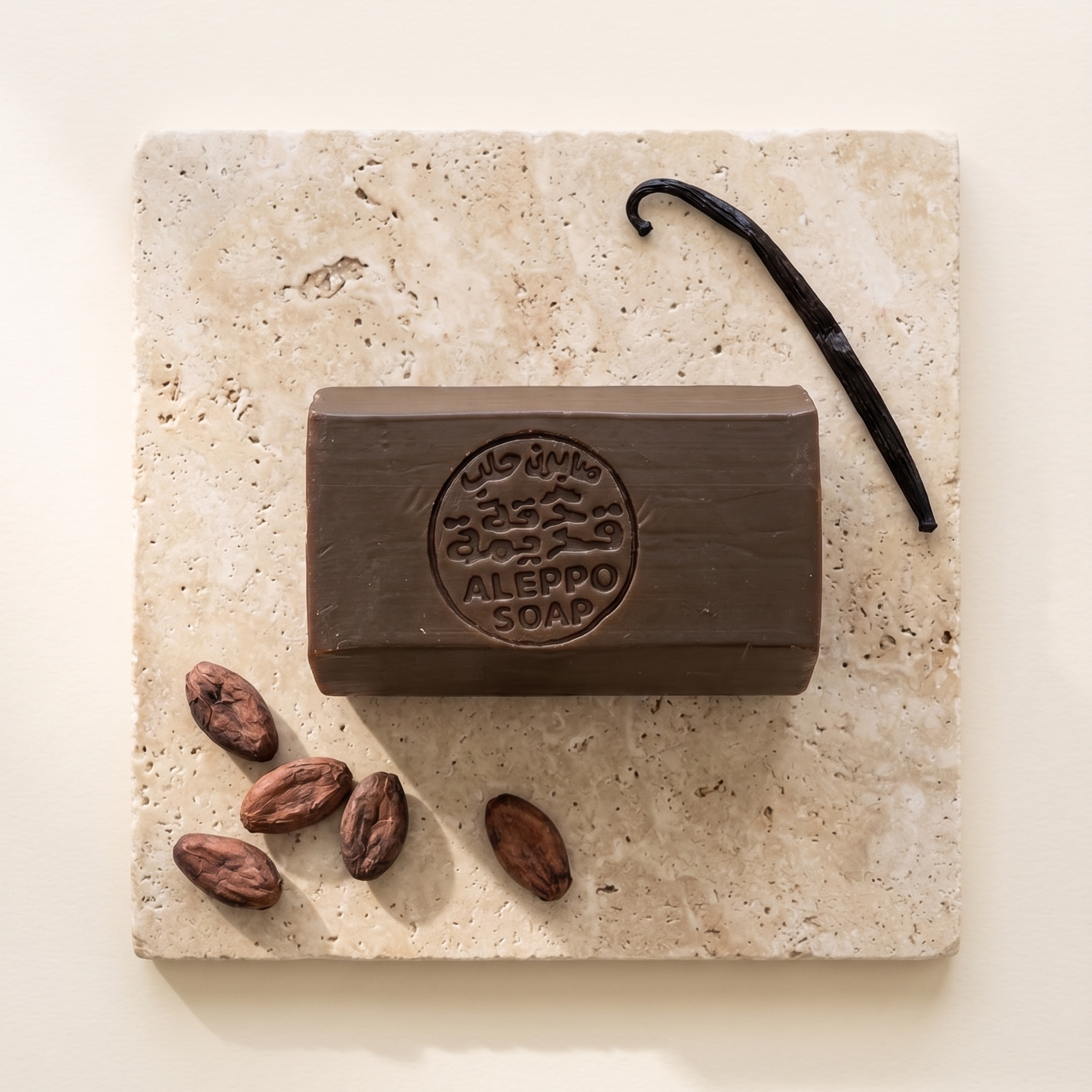 A rectangular bar of dark brown soap labeled 'MIO VINO ALEPPO SOAP' sits on a light beige stone slab, styled with cocoa beans and a vanilla bean on a warm cream background.