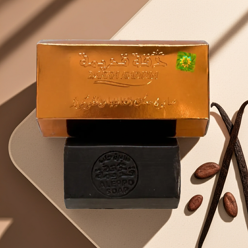 A bar of soap product shown with its metallic gold box on a beige surface. The box has Arabic and English branding text, including 'Mehmet Alimurad' and 'Almelo Soap', plus a small green flower logo.