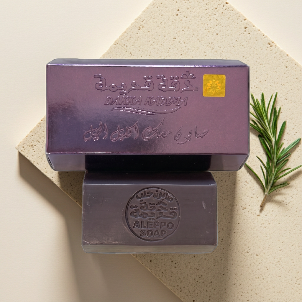 Two rectangular bars of soap stacked vertically, with the top bar showing its lid or outer packaging. The soap has a metallic lavender-purple finish with embossed Arabic calligraphy and English text reading 'ALERPPO SOAP'.