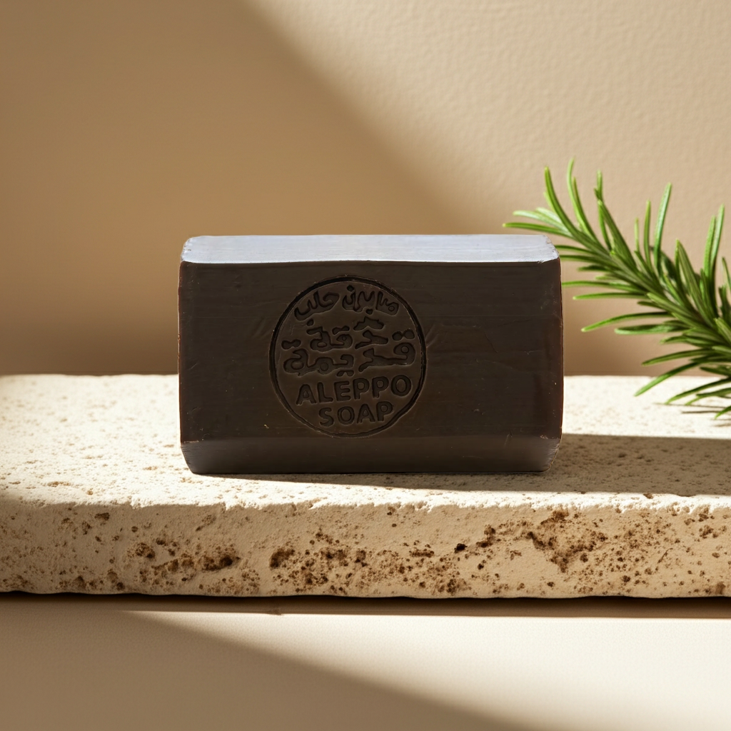 A rectangular bar of Aleppo Soap in a dark brown color, embossed with a circular logo, resting on a textured beige stone slab with a green pine branch in the background.
