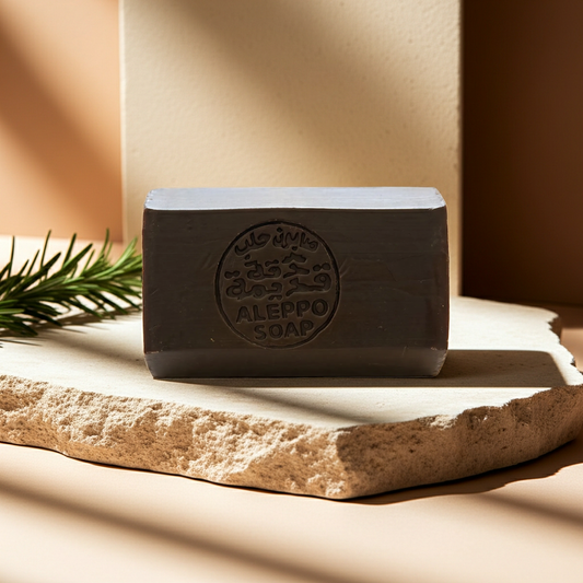 Rosemary Musk Aleppo Soap by Dakka Kadima — dark charcoal-grey bar of Aleppo soap on a textured stone slab, with a pine branch